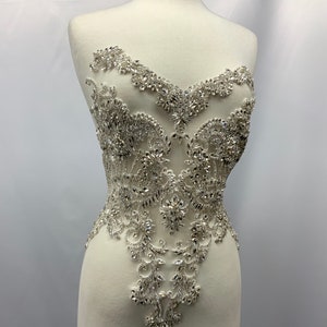 May include: A silver rhinestone applique with an intricate floral design. The applique is displayed on a white dress form. The design features a variety of shapes and sizes of rhinestones, creating a sparkling effect. The applique is likely for embellishing garments.