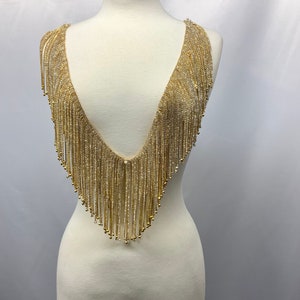 May include: Gold beaded fringe trim with a V-neck shape. The fringe is made of small, round beads that are strung together on a thin wire.