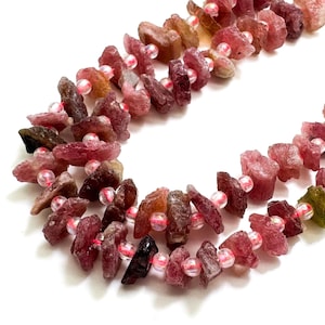 May include: Strands of raw, pink tourmaline gemstone beads. The beads are a mix of pink, red, and brown hues, with small, clear beads interspersed. The rough-cut stones are strung together, creating a natural and earthy aesthetic.