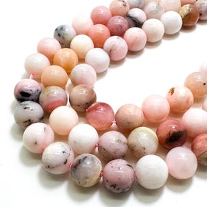 May include: Strands of round gemstone beads in shades of pink, peach, white, and brown. The beads have a polished, smooth surface and are strung together on a pink string. The beads are likely used for jewelry making.