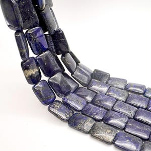 May include: A strand of rectangular lapis lazuli beads featuring a deep blue color with natural gold-toned pyrite inclusions. The beads are polished to a smooth finish and arranged in a curved line, showcasing their consistent shape and texture.
