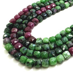 May include: Strands of faceted gemstone beads in shades of green and deep red. The beads are square-shaped and strung together, creating a textured and colorful display. The gemstones have a natural, earthy appearance.
