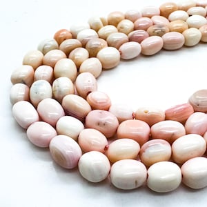 May include: Strands of oval-shaped beads in shades of pink, peach, and cream. The beads have a smooth, polished surface and are strung together, creating a necklace or bracelet design. The beads vary in color and size.