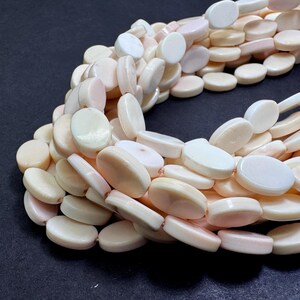 May include: Strands of oval-shaped, off-white beads with a subtle pink hue. The beads are smooth and appear to be strung together, creating a necklace or bracelet design. The beads are arranged on a dark surface.