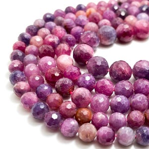 May include: Strands of faceted gemstone beads in shades of pink, purple, and mauve. The beads are round and have a textured, multi-faceted surface. The beads are strung together on a thin, pink string, and are suitable for jewelry making.