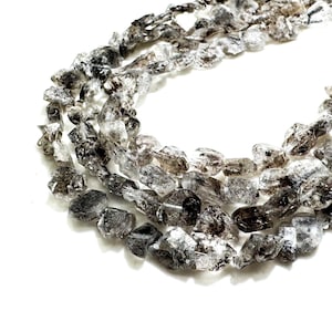Puede incluir: Una hebra de cuentas de cristal de cuarzo ahumado. Las cuentas son facetadas y tienen un aspecto áspero y natural. Las cuentas son de color gris claro con un tono ahumado.
