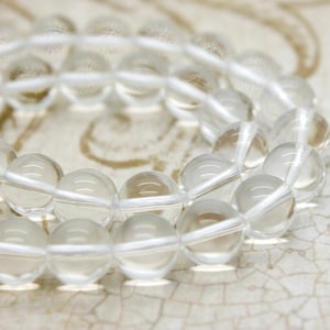 May include: A strand of clear quartz beads, strung on a white cord. The beads are round and smooth, and they are arranged in a loose, overlapping pattern.