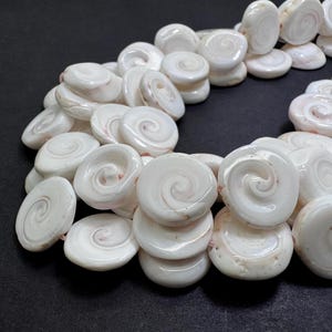 May include: Close-up of a strand of white shell beads. Each bead is a flat, circular shape with a spiral design in the center. The beads are strung together, creating a necklace or bracelet.