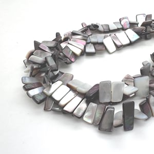 Puede incluir: Un collar hecho de cuentas rectangulares de nácar iridiscente. Las cuentas son en tonos grises, blancos y morados. El collar se exhibe sobre una superficie blanca. Las cuentas están ensartadas para formar un collar.