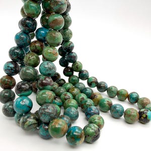 May include: A strand of round, polished beads featuring a variegated pattern of deep teal, forest green, and dark brown earthy tones. The beads are strung together in a loose, flowing arrangement, showcasing natural color variations and textures.