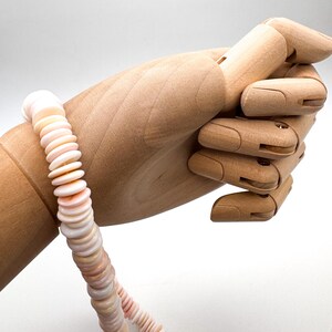 Op de afbeelding: Een ketting gemaakt van platte, ronde, lichtroze en witte schelpen. De ketting wordt getoond door een houten handmannequin tegen een witte achtergrond. De schelpen zijn aan elkaar geregen tot een doorlopende streng.
