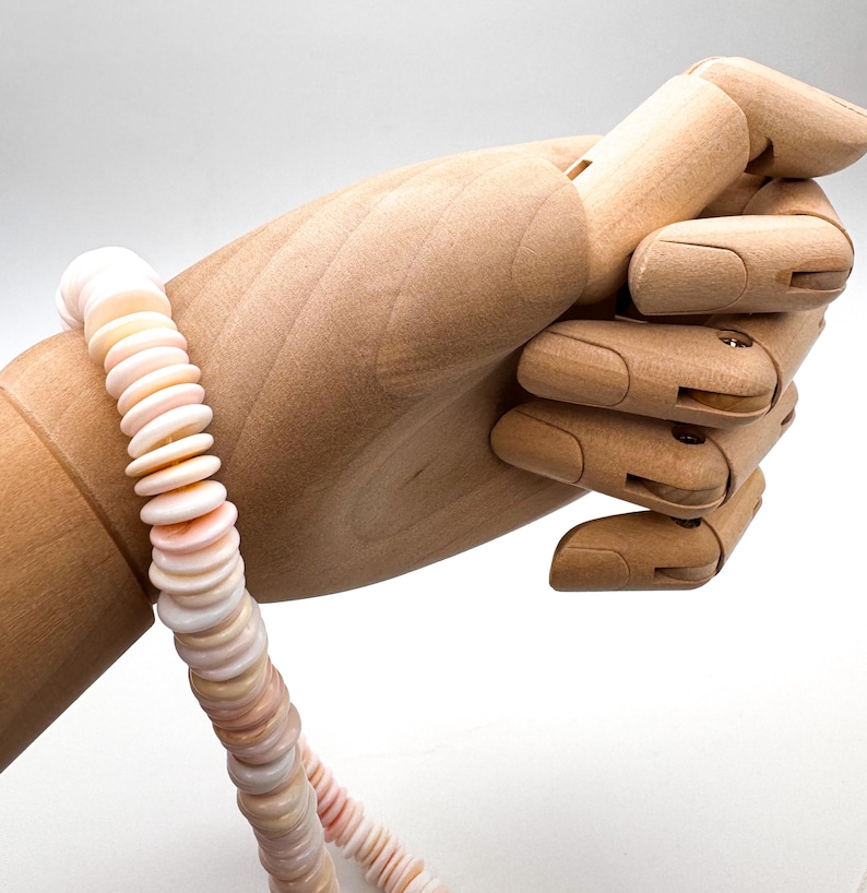 Op de afbeelding: Een ketting gemaakt van platte, ronde, lichtroze en witte schelpen. De ketting wordt getoond door een houten handmannequin tegen een witte achtergrond. De schelpen zijn aan elkaar geregen tot een doorlopende streng.