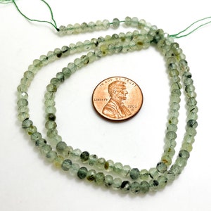 May include: A strand of green and black faceted gemstone beads, approximately 4mm in size, with a US penny for scale.
