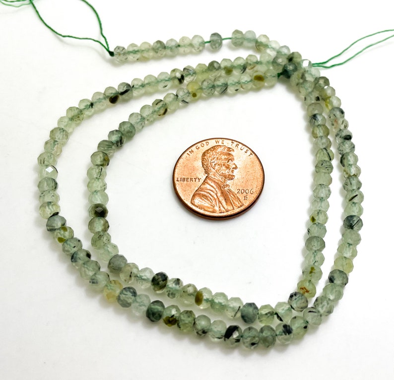 May include: A strand of green and black faceted gemstone beads, approximately 4mm in size, with a US penny for scale.