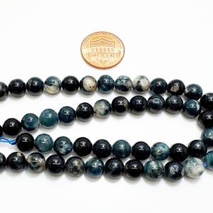 May include: Two strands of round, dark blue beads with a mix of solid and marbled textures. A copper penny is placed above the beads for size comparison. The beads are strung on blue thread, suitable for jewellery making.
