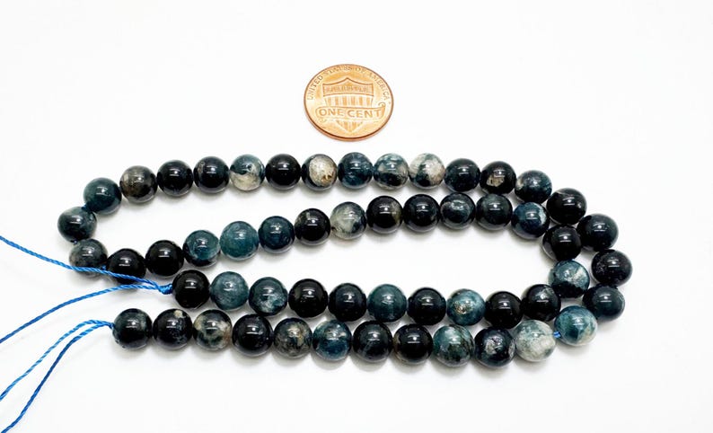 May include: Two strands of round, dark blue beads with a mix of solid and marbled textures. A copper penny is placed above the beads for size comparison. The beads are strung on blue thread, suitable for jewellery making.