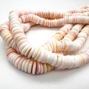 May include: Strands of pink and white shell beads. The beads are flat, disc-shaped, and strung together, creating a layered effect. The color palette includes soft pinks, creamy whites, and subtle yellow hues. The beads are likely used for jewelry making.