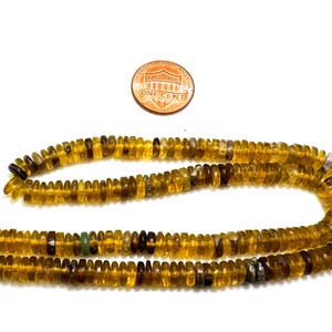 May include: Strands of amber-colored gemstone beads with a penny for size comparison. The beads are small, flat, and disc-shaped, with variations in color from light yellow to dark brown. The beads are strung on a yellow thread.