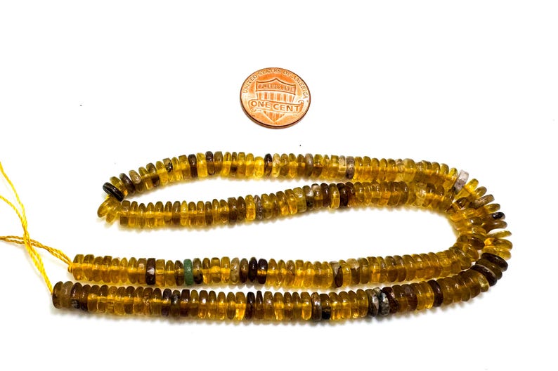 May include: Strands of amber-colored gemstone beads with a penny for size comparison. The beads are small, flat, and disc-shaped, with variations in color from light yellow to dark brown. The beads are strung on a yellow thread.