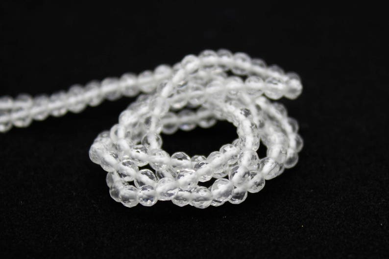 May include: A strand of clear, faceted beads, likely gemstones, is coiled on a black surface. The beads are small and round, reflecting light, suggesting a delicate jewelry item. The overall aesthetic is simple and elegant.