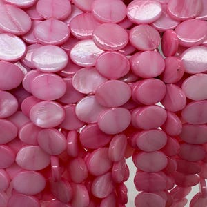 May include: Close-up of a strand of pink, iridescent, flat, round beads. The beads are strung together, creating a cascading effect. The beads have a glossy, polished finish and vary in shades of pink.