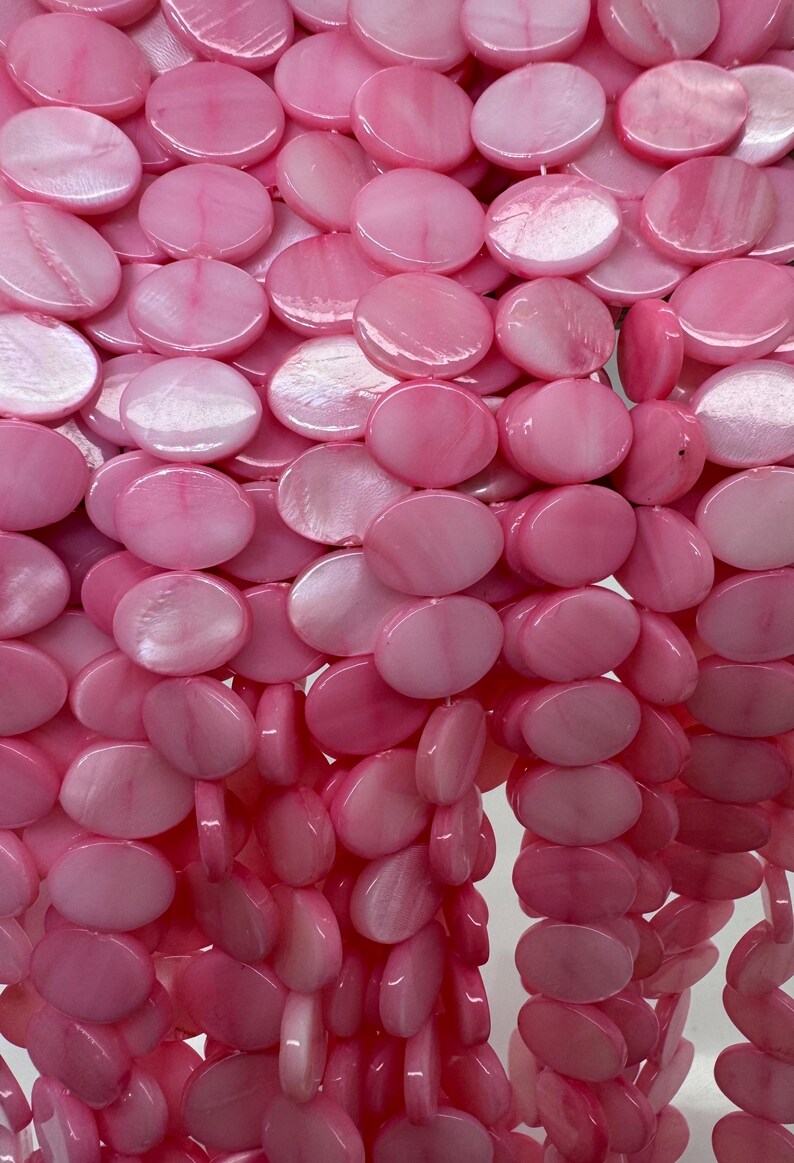 May include: Close-up of a strand of pink, iridescent, flat, round beads. The beads are strung together, creating a cascading effect. The beads have a glossy, polished finish and vary in shades of pink.