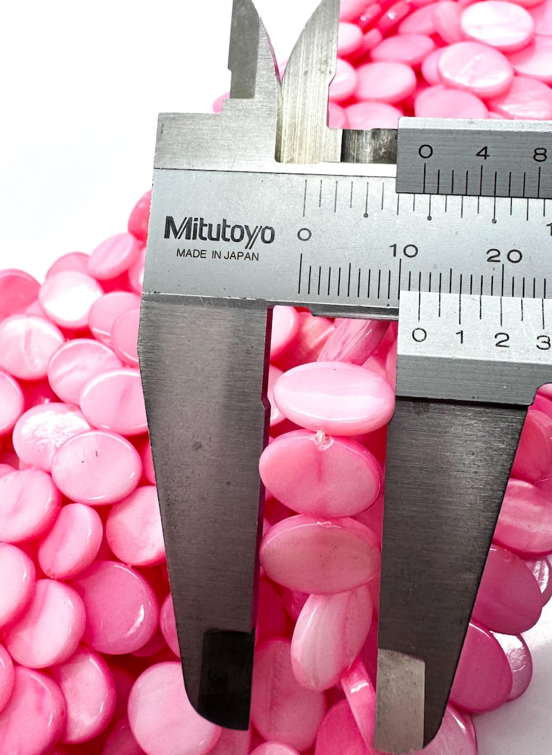May include: Close-up of pink, oval-shaped beads. A Mitutoyo caliper, made in Japan, measures the beads. The beads are strung together, and the caliper shows a measurement of approximately 0.3 inches.
