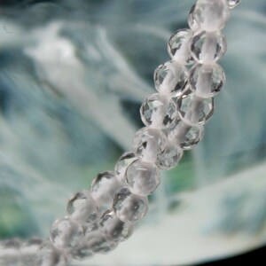 May include: Close-up of a strand of clear, faceted beads on a white string. The beads are round and reflect light, creating a sparkling effect. The background is a blurred mix of blue, green, and white, suggesting an abstract or natural theme.