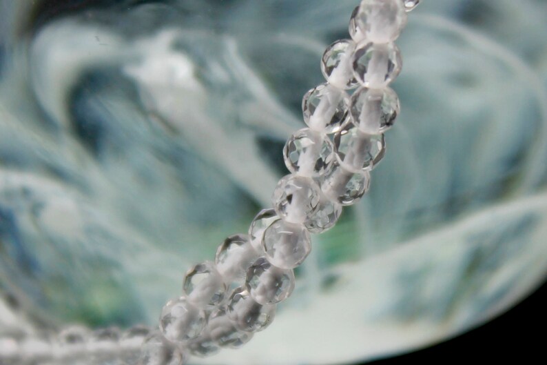 May include: Close-up of a strand of clear, faceted beads on a white string. The beads are round and reflect light, creating a sparkling effect. The background is a blurred mix of blue, green, and white, suggesting an abstract or natural theme.
