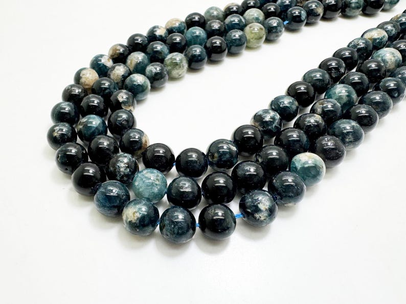 May include: Strands of polished, round beads in shades of dark blue, teal, and black. The beads have a glossy finish and are strung together on a blue cord. The beads are likely used for jewelry making.