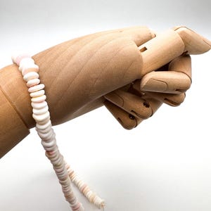 Op de afbeelding: Een delicate ketting gemaakt van kleine, platte, witte en roze schelpkralen. De kralen zijn aan elkaar geregen tot een doorlopende streng. De ketting wordt getoond op een houten handmodel, waarbij het ontwerp en de textuur worden benadrukt.