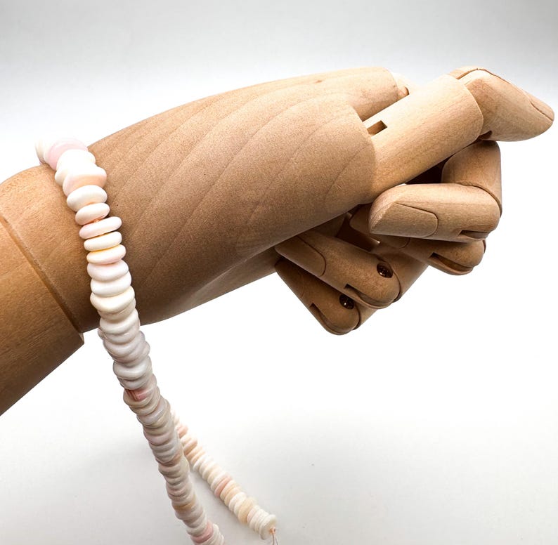 Op de afbeelding: Een delicate ketting gemaakt van kleine, platte, witte en roze schelpkralen. De kralen zijn aan elkaar geregen tot een doorlopende streng. De ketting wordt getoond op een houten handmodel, waarbij het ontwerp en de textuur worden benadrukt.