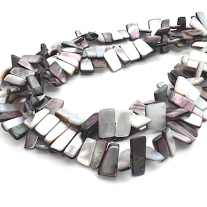 Può includere: Filo di perline rettangolari in madreperla nei toni del grigio, nero e bianco. Le perline sono disposte in un motivo fluido, creando un effetto strutturato e iridescente. Ideale per la creazione di gioielli.