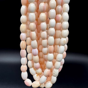 May include: A collection of multiple strands of smooth, oval-shaped beads in varying shades of pale pink, cream, and soft peach. The beads have a polished, semi-translucent appearance with subtle natural banding and variations in color throughout.