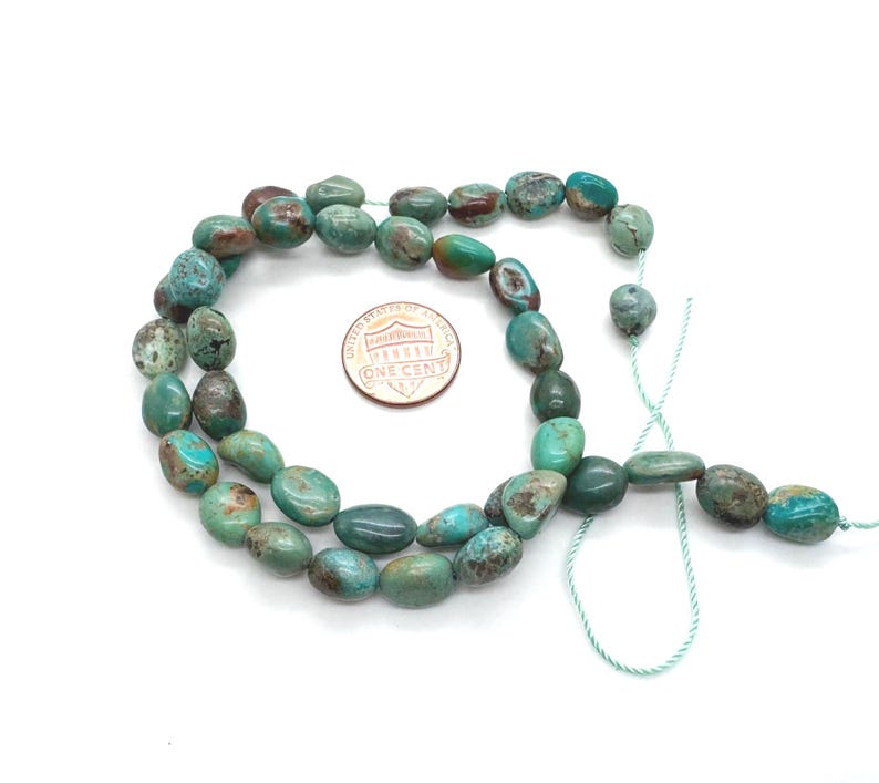 May include: Strand of turquoise-colored gemstone beads with a mix of blue, green, and brown hues. The beads are oval-shaped and strung on a light green cord. A US one-cent coin is next to the beads for size comparison.