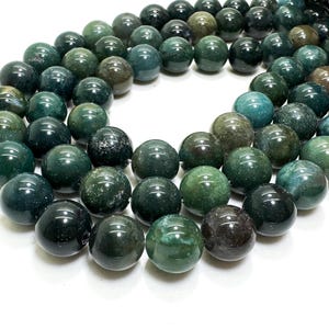 May include: A strand of polished, round beads in varying shades of green, teal, and dark brown. The beads have a smooth, glossy finish and are arranged in a curved line against a white background. The beads appear to be approximately 10mm in diameter.
