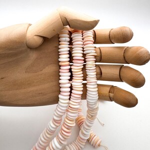 Op de afbeelding: Strengen roze en witte schelpkralen worden vastgehouden door een houten hand. De kralen zijn plat en rond, geregen om een delicate, natuurlijke uitstraling te cre&euml;ren. Het kleurenpalet is zacht, met variaties van roze en wit.