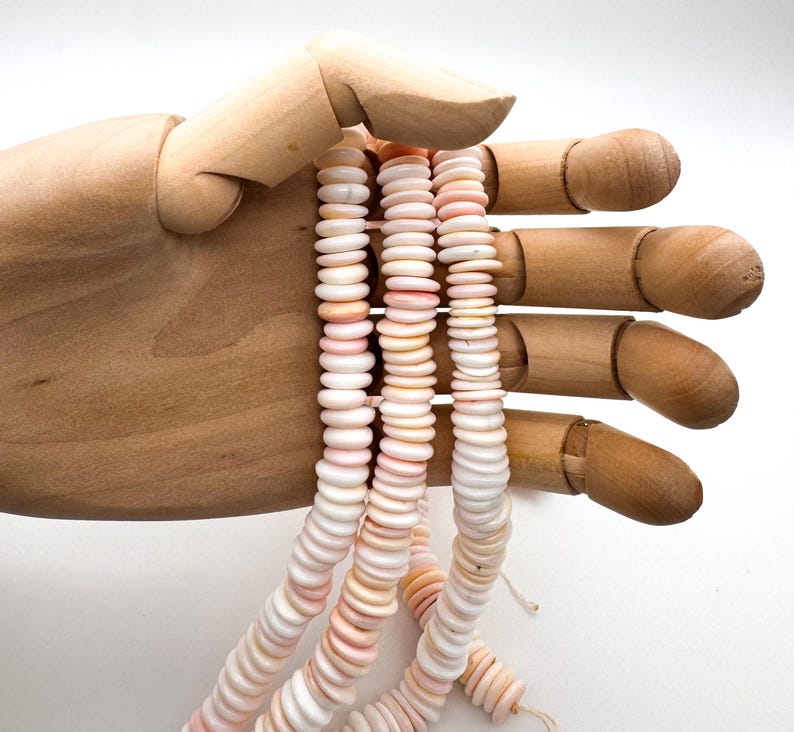 Op de afbeelding: Strengen roze en witte schelpkralen worden vastgehouden door een houten hand. De kralen zijn plat en rond, geregen om een delicate, natuurlijke uitstraling te cre&euml;ren. Het kleurenpalet is zacht, met variaties van roze en wit.