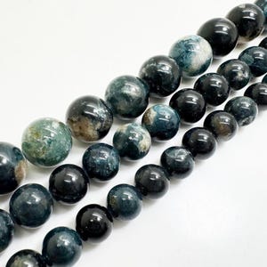 May include: A selection of polished gemstone beads in shades of blue, green, and brown. The beads are round and have a glossy finish, suitable for jewellery making. The beads are arranged diagonally against a white background.
