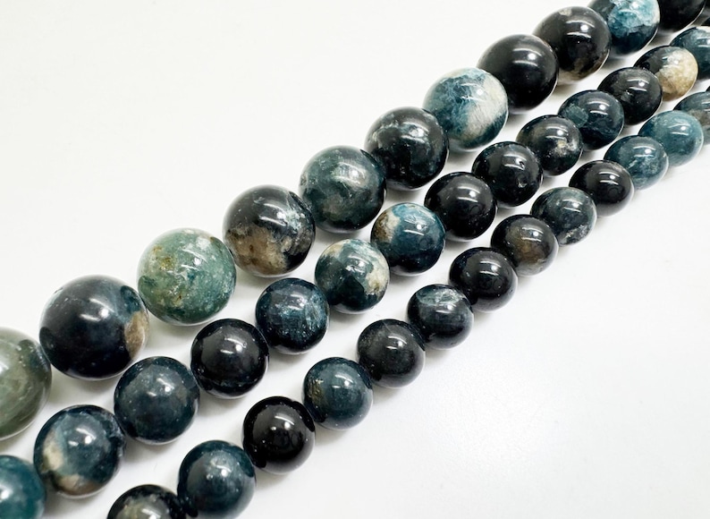 May include: A selection of polished gemstone beads in shades of blue, green, and brown. The beads are round and have a glossy finish, suitable for jewellery making. The beads are arranged diagonally against a white background.