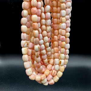 May include: Strands of pink and cream-colored beads. The beads are a mix of rounded and rectangular shapes, with variations in color and pattern. The beads are strung together, creating a cascading effect against a dark background.