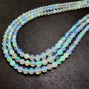 May include: Strands of iridescent opal beads. The beads are round and vary in size, with a milky white base color and flashes of blue, green, and yellow. The beads are strung together, creating a necklace or bracelet.