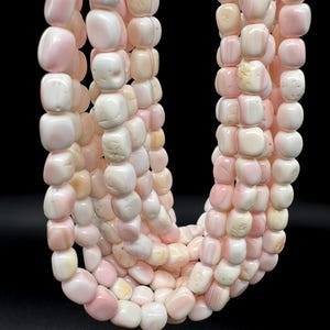 May include: A multi-strand necklace composed of numerous smooth, rounded, cube-shaped beads in varying shades of pale pink, cream, and soft white. The beads feature natural textures and subtle color variations, arranged in several cascading loops.