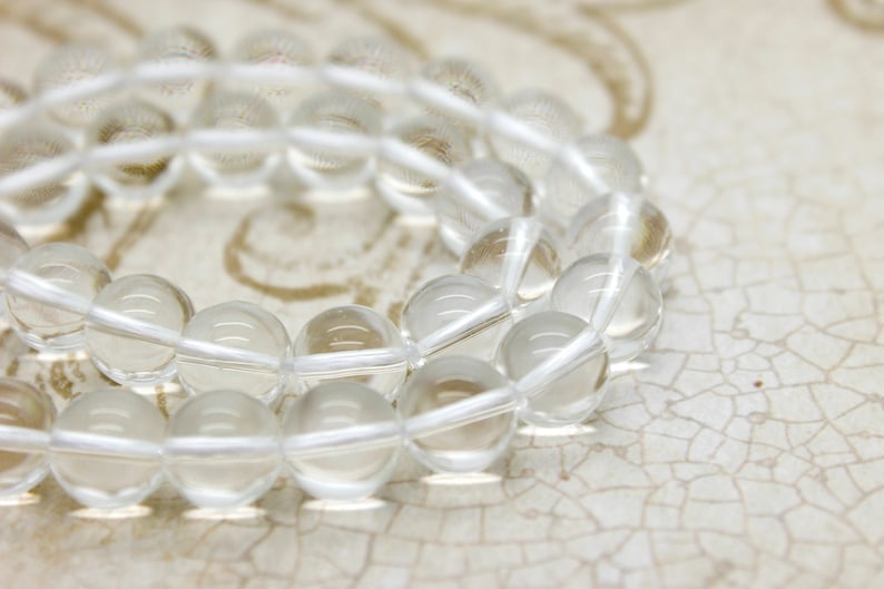 May include: A strand of clear quartz beads, strung on a white cord. The beads are round and smooth, and they are arranged in a loose, overlapping pattern.