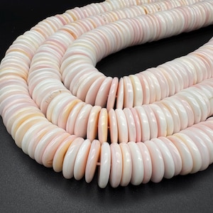 May include: A strand of circular, flat shell beads in varying shades of pale pink and white. The beads are arranged in a coiled, multi-layered formation, showcasing a smooth, polished texture and a consistent, uniform shape throughout the strand.