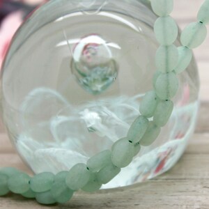 Puede incluir: Un collar de cuentas ovaladas de color verde claro. Las cuentas est&aacute;n ensartadas en un hilo fino de color verde oscuro. El collar est&aacute; delante de un globo de cristal transparente. El fondo est&aacute; borroso, con toques de rosa y morado.