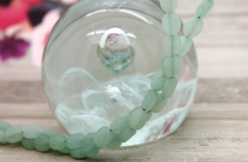 Puede incluir: Un collar de cuentas ovaladas de color verde claro. Las cuentas est&aacute;n ensartadas en un hilo fino de color verde oscuro. El collar est&aacute; delante de un globo de cristal transparente. El fondo est&aacute; borroso, con toques de rosa y morado.