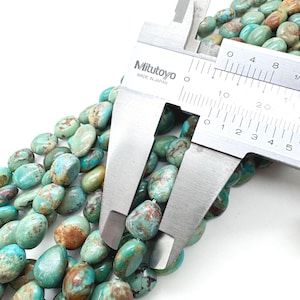 May include: Close-up of turquoise gemstone beads with brown matrix patterns. A Mitutoyo caliper, made in Japan, measures the beads. The beads are oval-shaped and have a polished finish. The color of the beads is a mix of blue-green and brown.