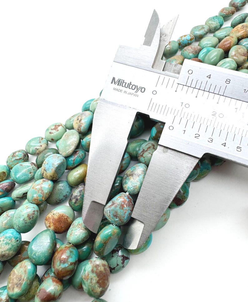 May include: Close-up of turquoise gemstone beads with brown matrix patterns. A Mitutoyo caliper, made in Japan, measures the beads. The beads are oval-shaped and have a polished finish. The color of the beads is a mix of blue-green and brown.