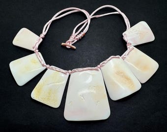 May include: A necklace featuring six rectangular, off-white pendants with subtle yellow and brown markings. The pendants are strung together with a pink cord, and the necklace has a matching pink clasp. The jewelry is displayed on a black surface.